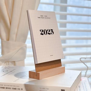 May include: A white desktop calendar with a wooden stand. The calendar is for the years 2025 and 2026. The front of the calendar has the year 2025 printed in large black letters.