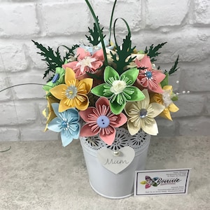 May include: A colourful paper flower bouquet in a white metal bucket. The flowers are in pastel shades of pink, yellow, blue, and green, each with a button or gem centre. A heart-shaped tag with the word "Mum" is attached to the bucket.