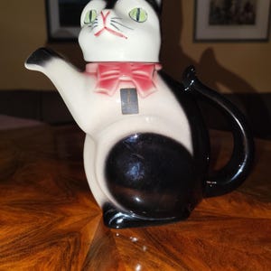 Könnte beinhalten: Eine Keramik-Teekanne in Form einer Katze, mit einem schwarz-weißen Körper, einer rosa Schleife und grünen Augen. Die Teekanne hat einen Griff und einen Ausguss und steht auf einer Holzoberfläche.