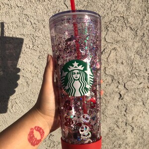 Puede incluir: Un vaso transparente de Starbucks lleno de purpurina rosa y morada y formas decorativas. El vaso tiene una base roja y una pajita roja. El logotipo de Starbucks es visible en la parte delantera del vaso.