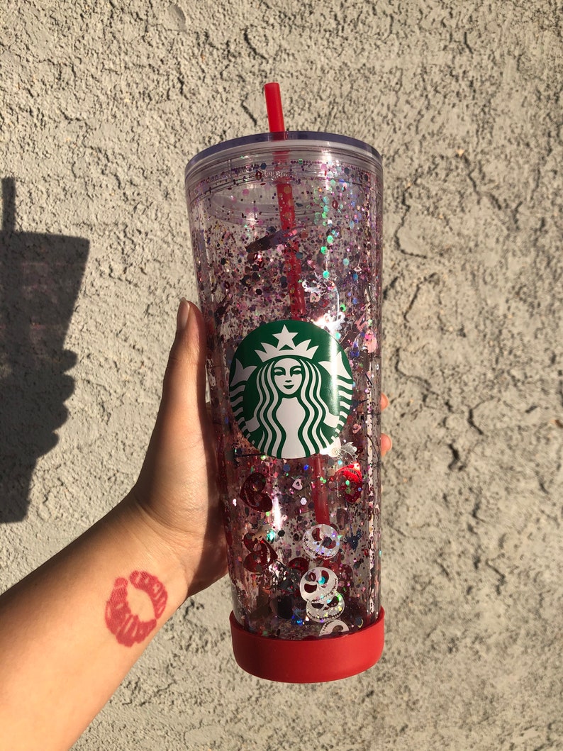 Puede incluir: Un vaso transparente de Starbucks lleno de purpurina rosa y morada y formas decorativas. El vaso tiene una base roja y una pajita roja. El logotipo de Starbucks es visible en la parte delantera del vaso.