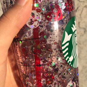 Puede incluir: Un vaso de pl&aacute;stico transparente lleno de purpurina roja, rosa e iridiscente y confeti en forma de coraz&oacute;n. Se ve una pajita roja en el interior. El logotipo de Starbucks est&aacute; en el lateral de la taza.