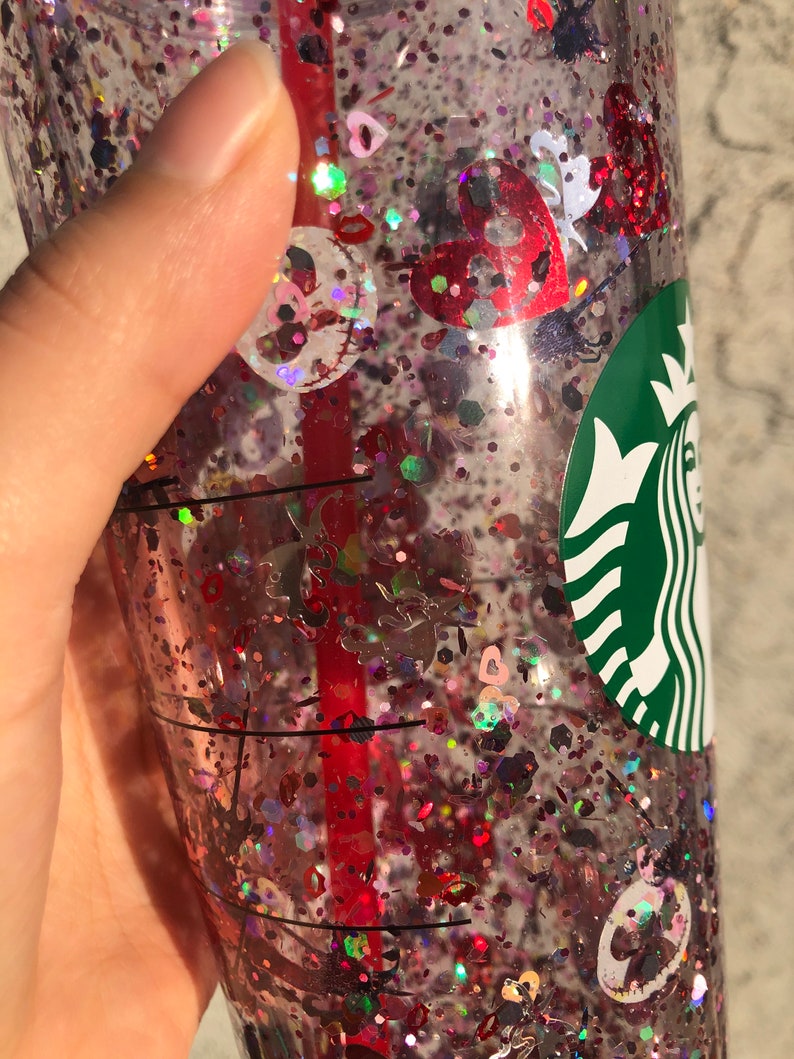Puede incluir: Un vaso de pl&aacute;stico transparente lleno de purpurina roja, rosa e iridiscente y confeti en forma de coraz&oacute;n. Se ve una pajita roja en el interior. El logotipo de Starbucks est&aacute; en el lateral de la taza.