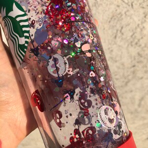Puede incluir: Un vaso transparente lleno de purpurina y confeti. El vaso tiene una base roja y un logotipo verde. La purpurina es roja, rosa e iridiscente, con confeti en forma de coraz&oacute;n y calavera. El dise&ntilde;o est&aacute; inspirado en una popular pel&iacute;cula de animaci&oacute;n.