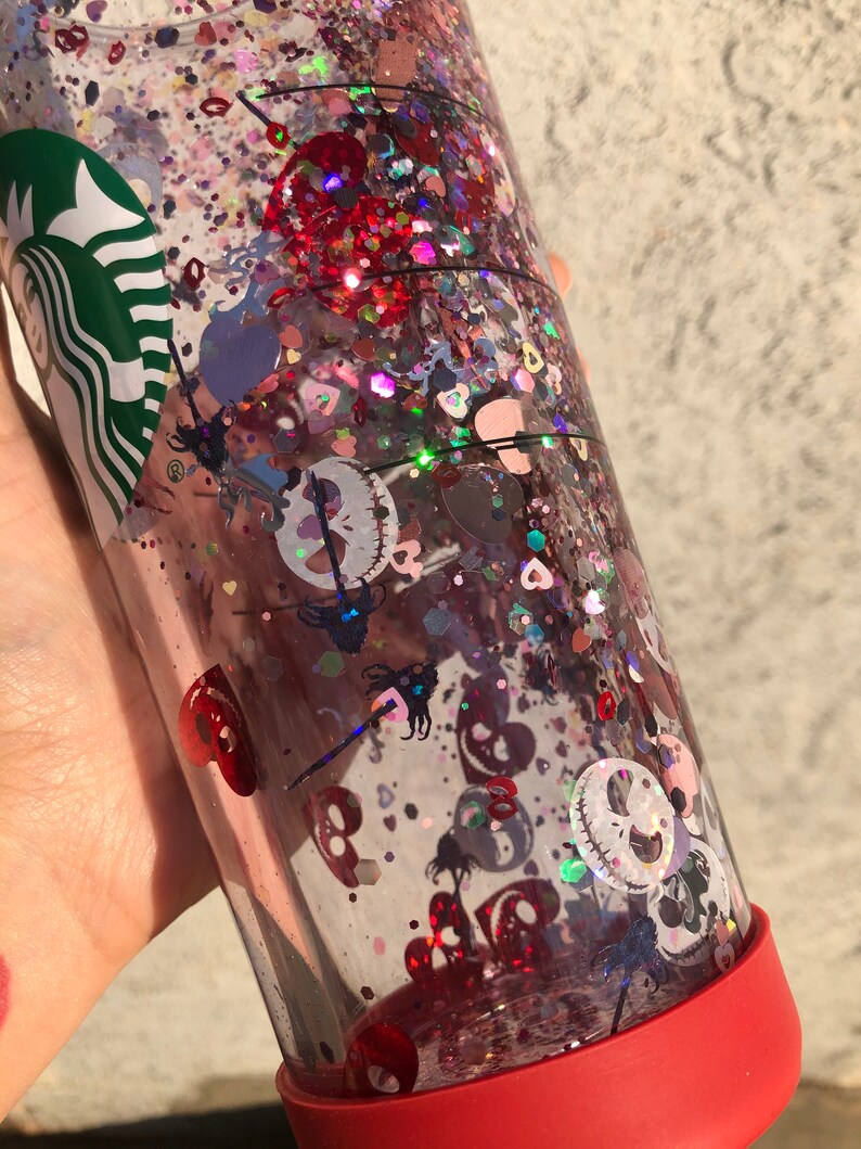 Puede incluir: Un vaso transparente lleno de purpurina y confeti. El vaso tiene una base roja y un logotipo verde. La purpurina es roja, rosa e iridiscente, con confeti en forma de coraz&oacute;n y calavera. El dise&ntilde;o est&aacute; inspirado en una popular pel&iacute;cula de animaci&oacute;n.