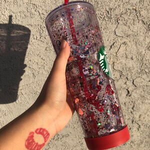 Puede incluir: Un vaso transparente de doble pared lleno de purpurina y confeti. El vaso tiene una base roja y una pajita roja. Un logotipo verde y blanco es visible en el lateral. El vaso se sostiene en una mano con un tatuaje de huella de labios rojos.