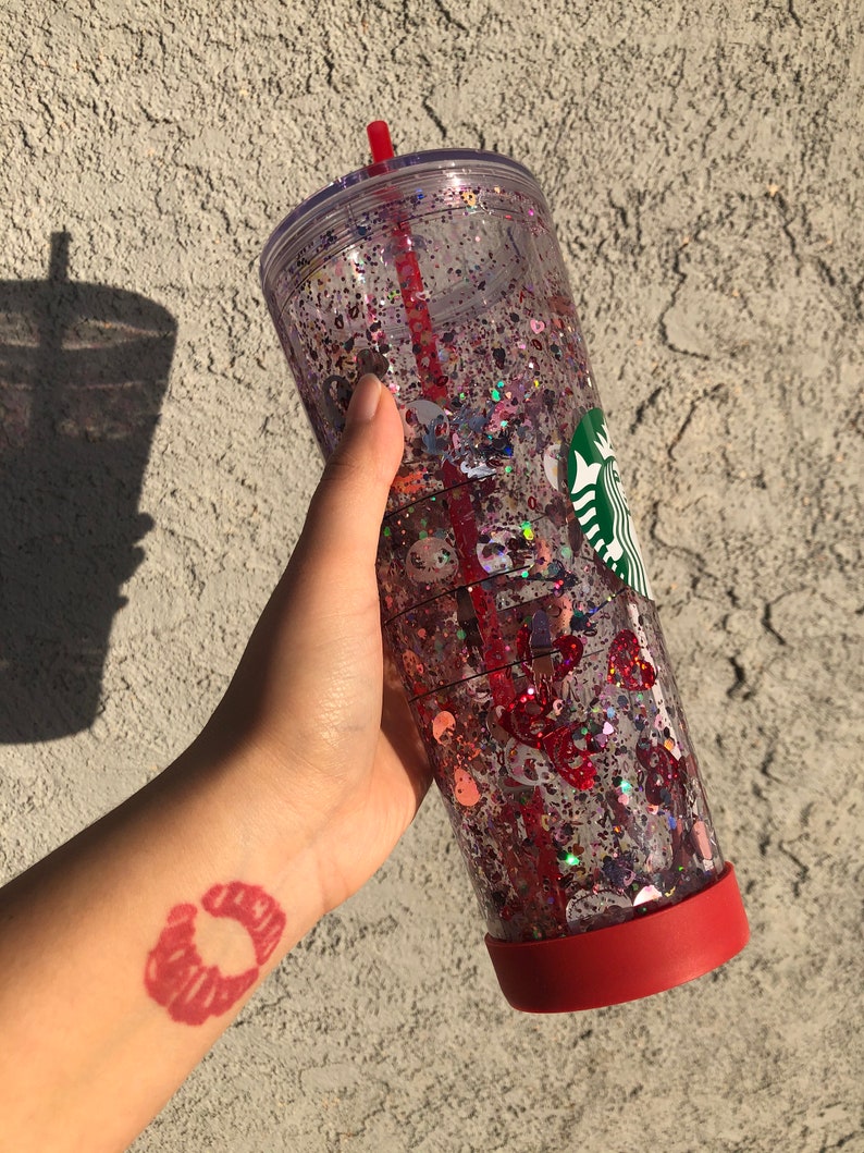 Puede incluir: Un vaso transparente de doble pared lleno de purpurina y confeti. El vaso tiene una base roja y una pajita roja. Un logotipo verde y blanco es visible en el lateral. El vaso se sostiene en una mano con un tatuaje de huella de labios rojos.
