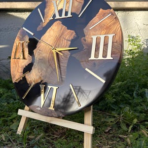 Peut inclure: Une horloge murale circulaire avec un centre en grain de bois naturel incrust&eacute; dans une r&eacute;sine noire brillante. Le cadran pr&eacute;sente des chiffres romains dor&eacute;s et des aiguilles fines dor&eacute;es, pos&eacute;e sur un support de pr&eacute;sentation en bois clair.