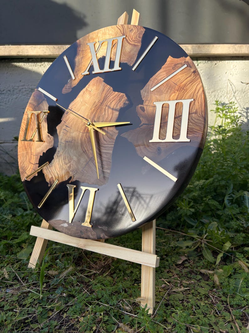 Peut inclure: Une horloge murale circulaire avec un centre en grain de bois naturel incrust&eacute; dans une r&eacute;sine noire brillante. Le cadran pr&eacute;sente des chiffres romains dor&eacute;s et des aiguilles fines dor&eacute;es, pos&eacute;e sur un support de pr&eacute;sentation en bois clair.