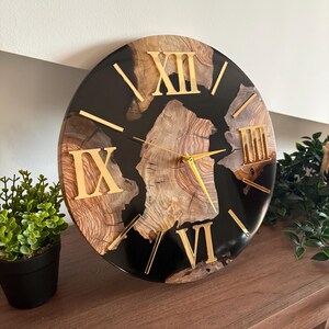Peut inclure: Une horloge murale ronde avec un centre en grain de bois naturel ench&acirc;ss&eacute; dans une bordure en r&eacute;sine noire lisse et brillante. Le cadran affiche des chiffres romains dor&eacute;s pour XII, IX, VI et III, avec des aiguilles dor&eacute;es fines.