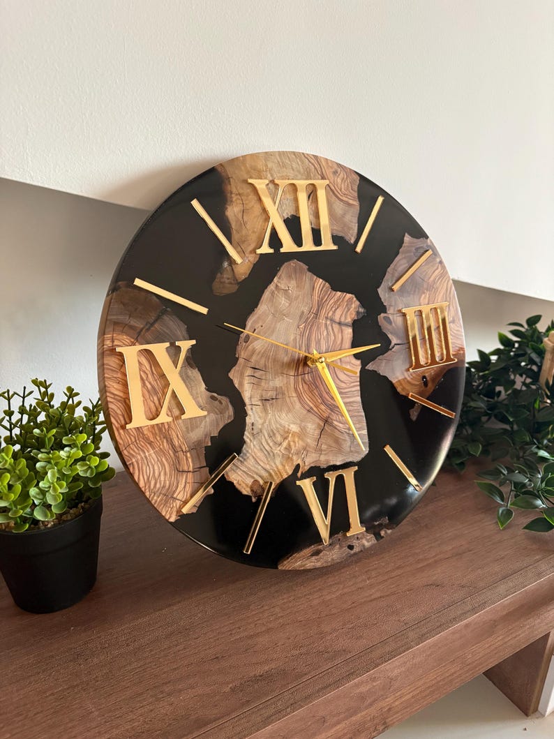 Peut inclure: Une horloge murale ronde avec un centre en grain de bois naturel ench&acirc;ss&eacute; dans une bordure en r&eacute;sine noire lisse et brillante. Le cadran affiche des chiffres romains dor&eacute;s pour XII, IX, VI et III, avec des aiguilles dor&eacute;es fines.