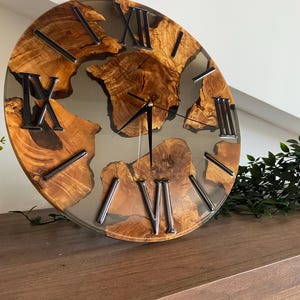 Peut inclure: Une horloge murale circulaire pr&eacute;sentant des sections de grain de bois brun clair naturel incrust&eacute;es dans une base en r&eacute;sine grise translucide et lisse. Le cadran affiche des chiffres romains noirs et de fines aiguilles en m&eacute;tal noir au centre, cr&eacute;ant un contraste entre les textures organiques du bois et le mat&eacute;riau moderne et &eacute;l&eacute;gant de la r&eacute;sine.