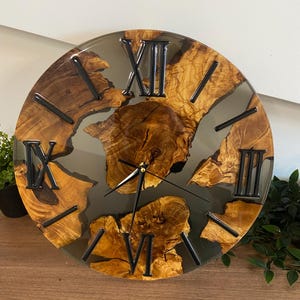 Peut inclure: Une horloge murale ronde compos&eacute;e d'une tranche de bois naturel &agrave; bord brut incrust&eacute;e dans de la r&eacute;sine gris fonc&eacute; translucide. Le cadran pr&eacute;sente des chiffres romains noirs et de fines aiguilles en m&eacute;tal noir sur le bois vein&eacute;.