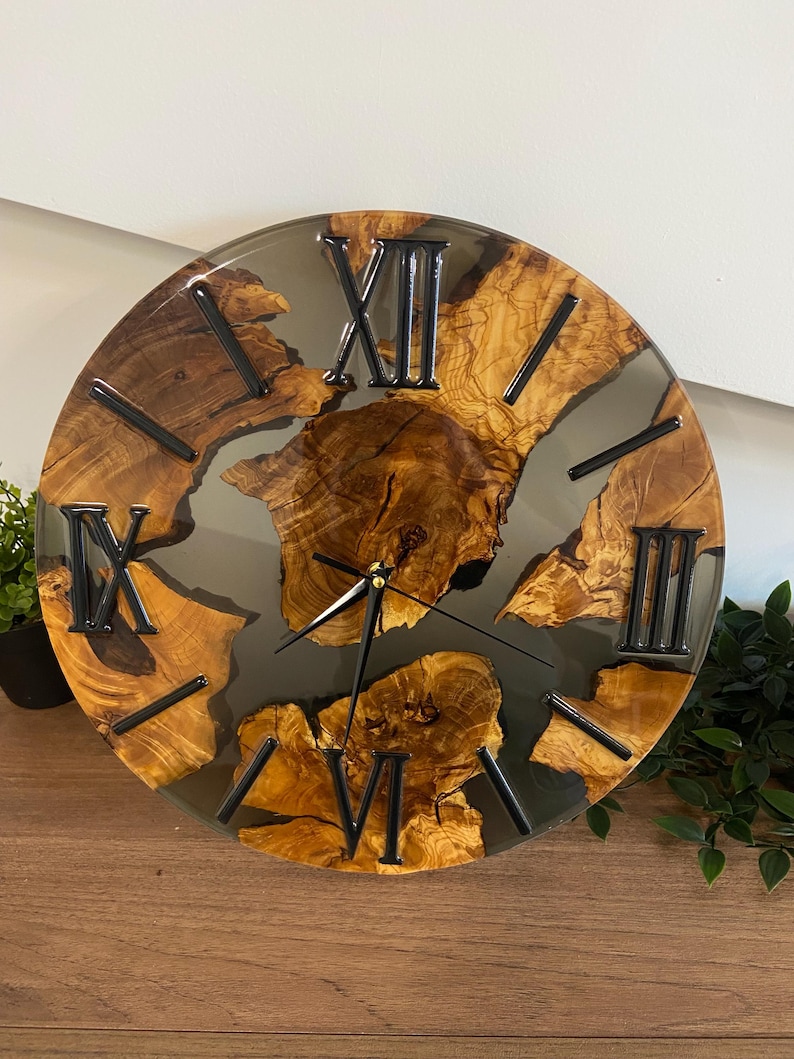 Peut inclure: Une horloge murale ronde compos&eacute;e d'une tranche de bois naturel &agrave; bord brut incrust&eacute;e dans de la r&eacute;sine gris fonc&eacute; translucide. Le cadran pr&eacute;sente des chiffres romains noirs et de fines aiguilles en m&eacute;tal noir sur le bois vein&eacute;.