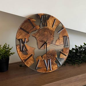 Peut inclure: Une horloge murale circulaire compos&eacute;e de sections de bois naturel incrust&eacute;es dans une base en r&eacute;sine grise translucide. Le cadran pr&eacute;sente des chiffres romains m&eacute;talliques sombres et de fines aiguilles noires, contrastant avec le bois organique et la r&eacute;sine lisse.