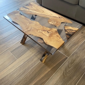 Op de afbeelding: Een rechthoekige salontafel met een helder harsmidden en natuurlijke houten randen. De tafel heeft een donker metalen frame en houten poten. Het hout heeft een lichtbruine kleur met donkerdere bruine nerfpatronen.