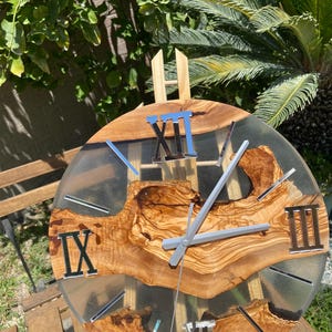 Peut inclure: Une horloge murale ronde compos&eacute;e d'une tranche de bois naturel &agrave; bord brut incrust&eacute;e dans de la r&eacute;sine translucide. Le cadran pr&eacute;sente des chiffres romains argent&eacute;s aux positions douze, trois, six et neuf heures, avec des aiguilles argent&eacute;es.