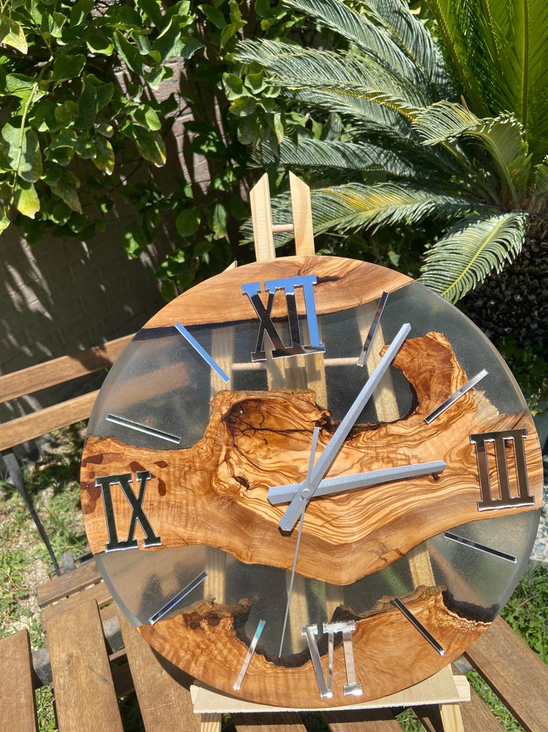 Peut inclure: Une horloge murale ronde compos&eacute;e d'une tranche de bois naturel &agrave; bord brut incrust&eacute;e dans de la r&eacute;sine translucide. Le cadran pr&eacute;sente des chiffres romains argent&eacute;s aux positions douze, trois, six et neuf heures, avec des aiguilles argent&eacute;es.