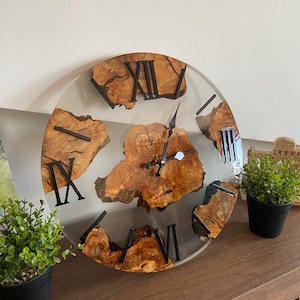 Peut inclure: Une horloge murale ronde compos&eacute;e d'une tranche de bois naturel au centre et de segments incrust&eacute;s dans de la r&eacute;sine transparente. Le cadran pr&eacute;sente des chiffres romains noirs et des aiguilles en m&eacute;tal noir sur le bois vein&eacute;.