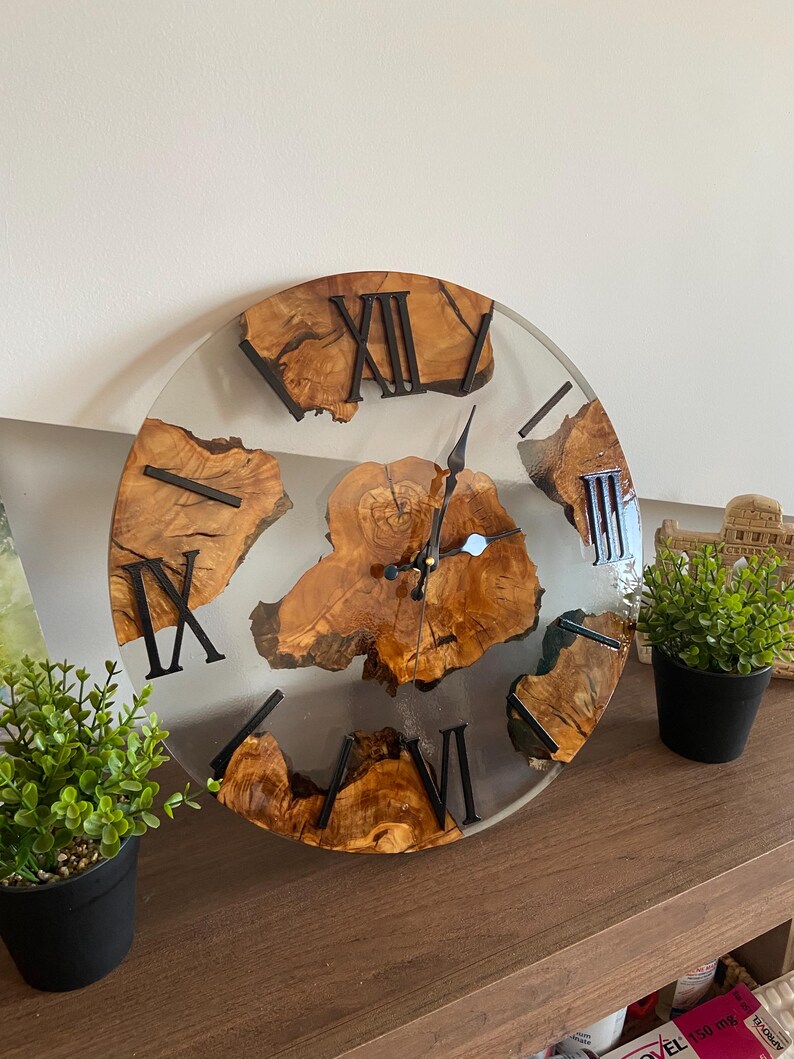 Peut inclure: Une horloge murale ronde compos&eacute;e d'une tranche de bois naturel au centre et de segments incrust&eacute;s dans de la r&eacute;sine transparente. Le cadran pr&eacute;sente des chiffres romains noirs et des aiguilles en m&eacute;tal noir sur le bois vein&eacute;.