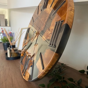 Peut inclure: Une horloge murale ronde pr&eacute;sentant une surface polie en loupe de bois naturel combin&eacute;e &agrave; de la r&eacute;sine sombre et translucide. Le cadran affiche des chiffres romains noirs en relief et de fines aiguilles m&eacute;talliques, avec une finition brillante.