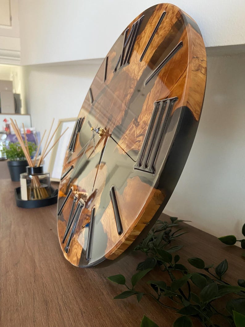 Peut inclure: Une horloge murale ronde pr&eacute;sentant une surface polie en loupe de bois naturel combin&eacute;e &agrave; de la r&eacute;sine sombre et translucide. Le cadran affiche des chiffres romains noirs en relief et de fines aiguilles m&eacute;talliques, avec une finition brillante.