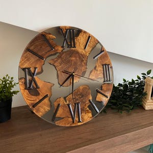 Peut inclure: Une horloge murale ronde compos&eacute;e de tranches de bois naturel incrust&eacute;es dans de la r&eacute;sine grise translucide. Le cadran pr&eacute;sente des chiffres romains noirs et de fines aiguilles noires. Le grain du bois est visible, cr&eacute;ant un contraste entre les bords organiques du bois et la r&eacute;sine lisse et semi-transparente.
