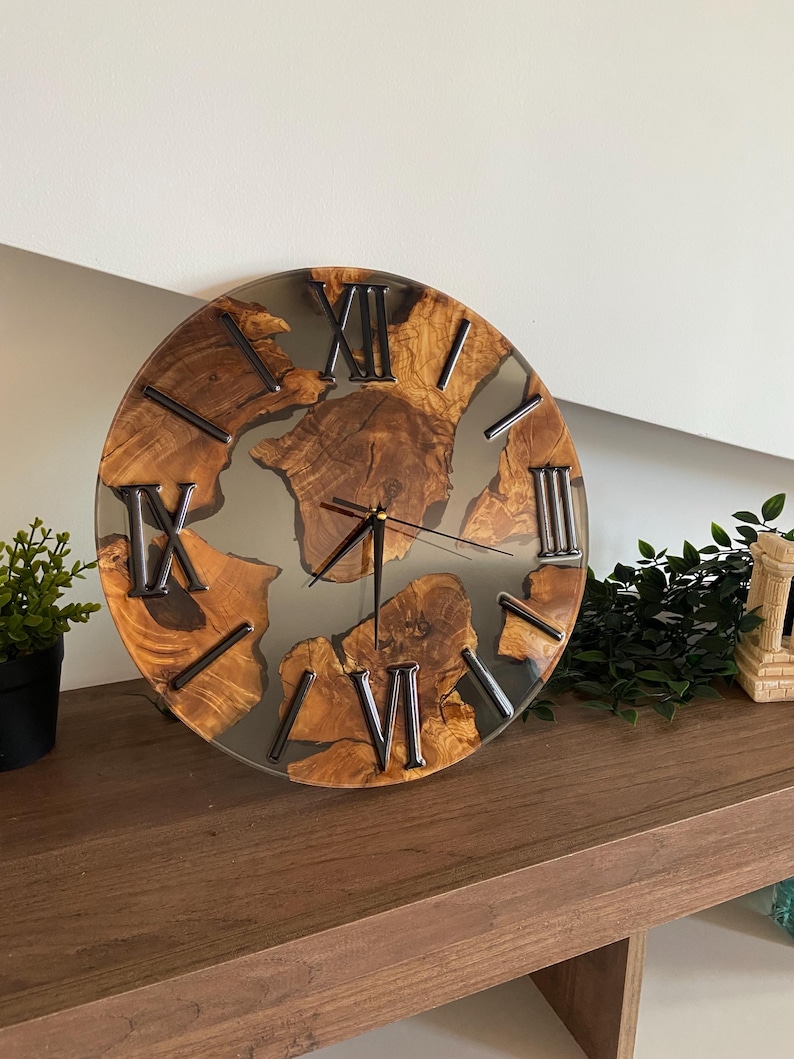 Peut inclure: Une horloge murale ronde compos&eacute;e de tranches de bois naturel incrust&eacute;es dans de la r&eacute;sine grise translucide. Le cadran pr&eacute;sente des chiffres romains noirs et de fines aiguilles noires. Le grain du bois est visible, cr&eacute;ant un contraste entre les bords organiques du bois et la r&eacute;sine lisse et semi-transparente.