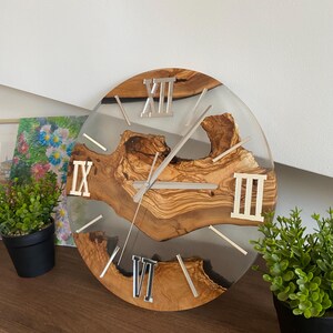 Peut inclure: Une horloge murale ronde compos&eacute;e d'une tranche de bois naturel &agrave; bord brut incrust&eacute;e dans de la r&eacute;sine transparente. Le cadran pr&eacute;sente des chiffres romains argent&eacute;s aux positions douze, trois, six et neuf heures, avec des aiguilles argent&eacute;es.