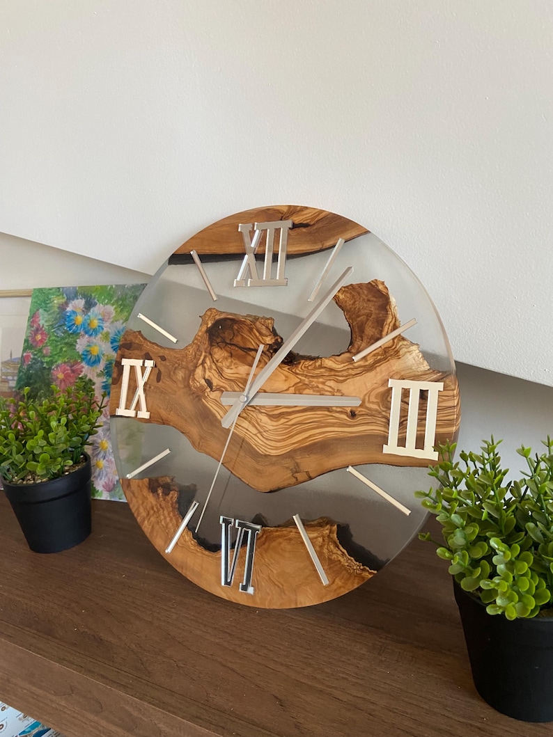 Peut inclure: Une horloge murale ronde compos&eacute;e d'une tranche de bois naturel &agrave; bord brut incrust&eacute;e dans de la r&eacute;sine transparente. Le cadran pr&eacute;sente des chiffres romains argent&eacute;s aux positions douze, trois, six et neuf heures, avec des aiguilles argent&eacute;es.