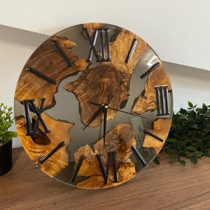 Peut inclure: Une horloge murale ronde compos&eacute;e de sections de bois naturel incrust&eacute;es dans de la r&eacute;sine grise translucide. Le cadran pr&eacute;sente des chiffres romains noirs et de fines aiguilles noires. Le bois est brun chaud avec des n&oelig;uds visibles.