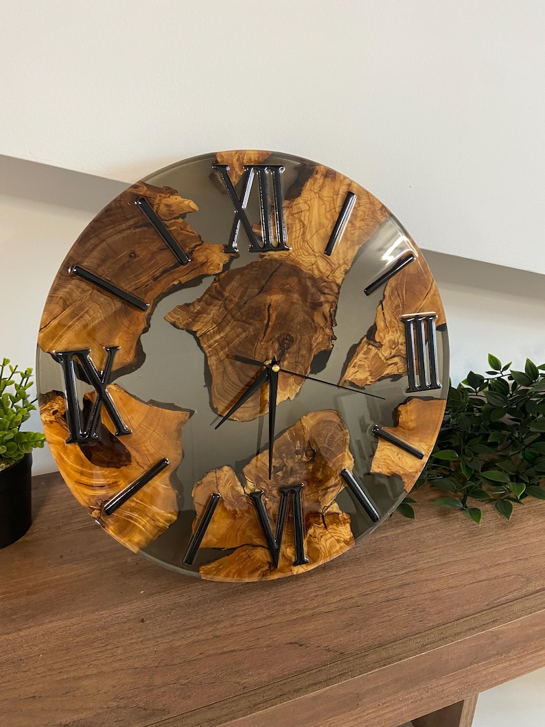 Peut inclure: Une horloge murale ronde compos&eacute;e de sections de bois naturel incrust&eacute;es dans de la r&eacute;sine grise translucide. Le cadran pr&eacute;sente des chiffres romains noirs et de fines aiguilles noires. Le bois est brun chaud avec des n&oelig;uds visibles.