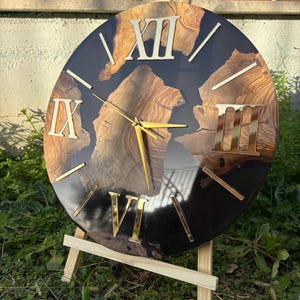 Peut inclure: Une horloge murale ronde compos&eacute;e d'une tranche de bois naturel aux motifs de grain marqu&eacute;s, incrust&eacute;e dans une base en r&eacute;sine noire brillante. Le cadran pr&eacute;sente des chiffres romains dor&eacute;s XII, IX, VI et III, ainsi que des aiguilles dor&eacute;es.