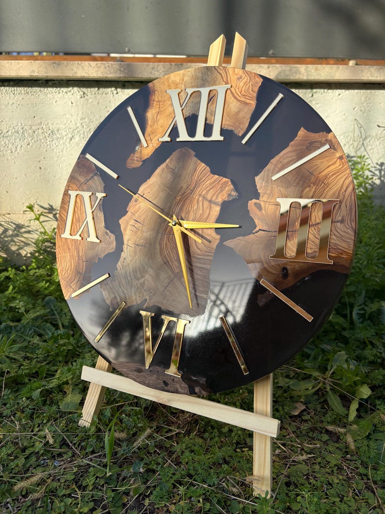 Peut inclure: Une horloge murale ronde compos&eacute;e d'une tranche de bois naturel aux motifs de grain marqu&eacute;s, incrust&eacute;e dans une base en r&eacute;sine noire brillante. Le cadran pr&eacute;sente des chiffres romains dor&eacute;s XII, IX, VI et III, ainsi que des aiguilles dor&eacute;es.