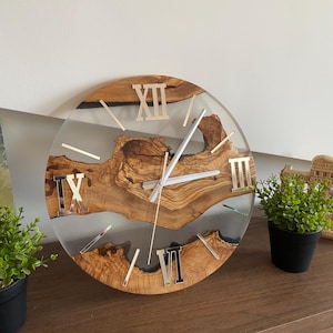 Peut inclure: Une horloge murale ronde compos&eacute;e d'une tranche de bois naturel &agrave; bord brut incrust&eacute;e dans de la r&eacute;sine transparente. Le cadran pr&eacute;sente des chiffres romains argent&eacute;s aux positions douze, neuf et six heures, avec des aiguilles argent&eacute;es.