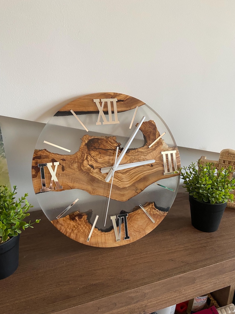Peut inclure: Une horloge murale ronde compos&eacute;e d'une tranche de bois naturel &agrave; bord brut incrust&eacute;e dans de la r&eacute;sine transparente. Le cadran pr&eacute;sente des chiffres romains argent&eacute;s aux positions douze, neuf et six heures, avec des aiguilles argent&eacute;es.