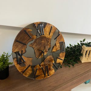Peut inclure: Une horloge murale ronde compos&eacute;e de segments de bois naturel incrust&eacute;s dans une r&eacute;sine grise translucide. Le cadran pr&eacute;sente des chiffres romains noirs et de fines aiguilles noires. Le grain du bois est visible sur les morceaux de bois.