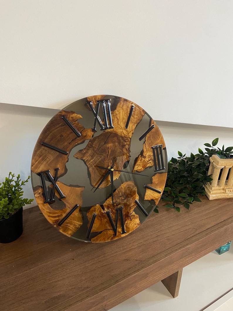 Peut inclure: Une horloge murale ronde compos&eacute;e de segments de bois naturel incrust&eacute;s dans une r&eacute;sine grise translucide. Le cadran pr&eacute;sente des chiffres romains noirs et de fines aiguilles noires. Le grain du bois est visible sur les morceaux de bois.