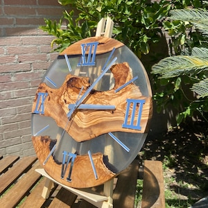 Peut inclure: Une horloge murale ronde compos&eacute;e d'une tranche de bois d'olivier naturel incrust&eacute;e dans de la r&eacute;sine &eacute;poxy transparente. Le cadran pr&eacute;sente des chiffres romains argent&eacute;s et des aiguilles assorties, soulignant le grain organique du bois.