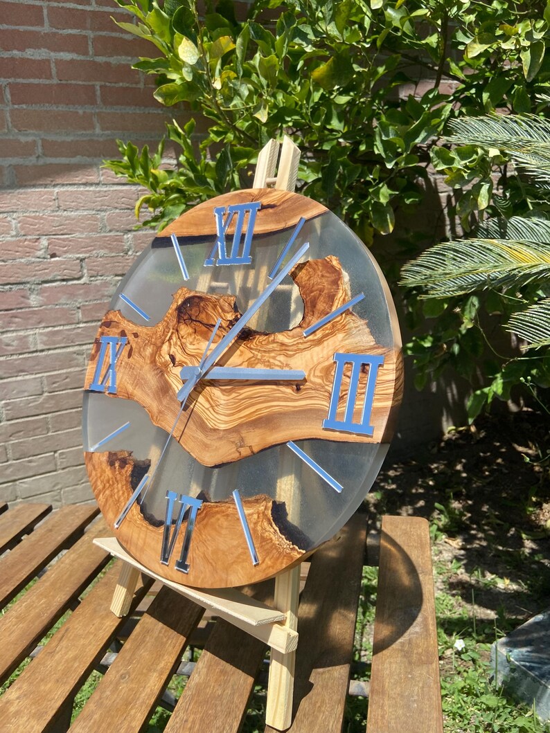 Peut inclure: Une horloge murale ronde compos&eacute;e d'une tranche de bois d'olivier naturel incrust&eacute;e dans de la r&eacute;sine &eacute;poxy transparente. Le cadran pr&eacute;sente des chiffres romains argent&eacute;s et des aiguilles assorties, soulignant le grain organique du bois.