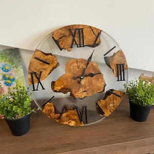 Peut inclure: Une horloge murale ronde compos&eacute;e d'une pi&egrave;ce centrale et de segments ext&eacute;rieurs en bois clair naturel au grain visible, incrust&eacute;s dans de la r&eacute;sine transparente. Elle poss&egrave;de des aiguilles en m&eacute;tal noir et des chiffres romains noirs.