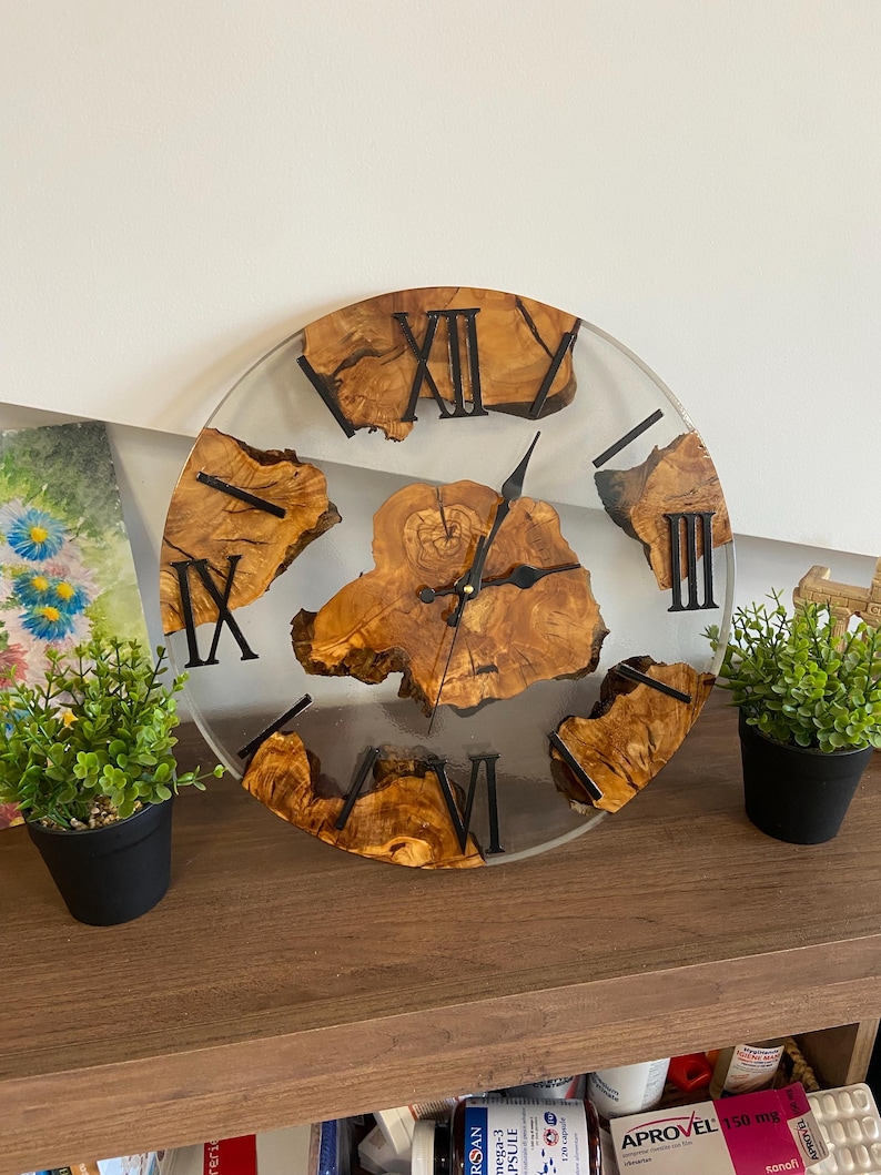 Peut inclure: Une horloge murale ronde compos&eacute;e d'une pi&egrave;ce centrale et de segments ext&eacute;rieurs en bois clair naturel au grain visible, incrust&eacute;s dans de la r&eacute;sine transparente. Elle poss&egrave;de des aiguilles en m&eacute;tal noir et des chiffres romains noirs.