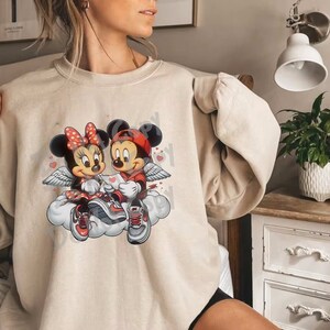 May include: Beige sweatshirt featuring a graphic of Mickey and Minnie Mouse as cupids, seated on a cloud. Minnie wears a red bow and dress, Mickey a red cap and jacket. Both have wings and sneakers. Hearts float around them.