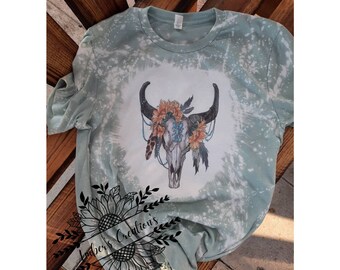 May include: A light green t-shirt with a bleached design. The shirt features a graphic of a bull skull adorned with flowers, feathers, and beads. The text "Amber's Creations" is printed on the shirt.