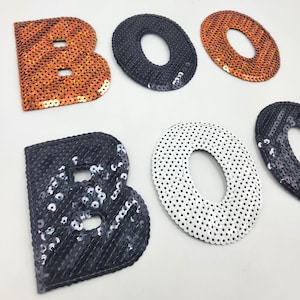May include: Sequined letters spelling "BOO". The letters are in orange, black, and white, with a sequined texture. The letters are likely for a Halloween or festive decoration.