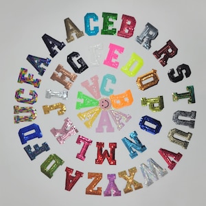 Peut inclure: Un assortiment de lettres de l'alphabet pailletées et colorées, disposées en cercle. Les lettres, de couleurs variées comme le bleu, le vert, le rose et l'argent, sont probablement destinées à l'artisanat.