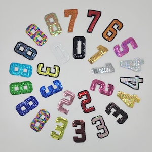 May include: A collection of sequined number patches in various colors, including gold, pink, blue, and green. The numbers 0 through 8 are displayed, each with a different color and sequin pattern. These patches are likely for clothing or craft projects.