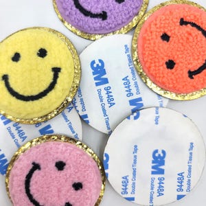 Peut inclure: Gros plan sur des écussons smiley colorés. Les écussons sont jaunes, violets, roses et oranges avec des sourires brodés noirs. Chaque écusson a une bordure dorée scintillante et se trouve à côté d'un support adhésif blanc.