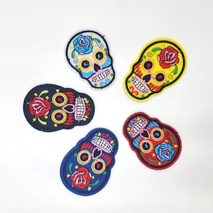 May include: Five colorful embroidered skull patches with floral accents. The patches feature a variety of colors, including blue, yellow, red, and black, with intricate details and stitching. The skulls are a Day of the Dead theme.