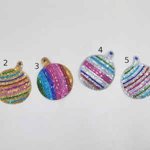 May include: Six sequined, round ornaments with a loop at the top. Each ornament features a different colour scheme of horizontal stripes. Colours include gold, pink, blue, silver, and purple. Suitable for holiday decorations.